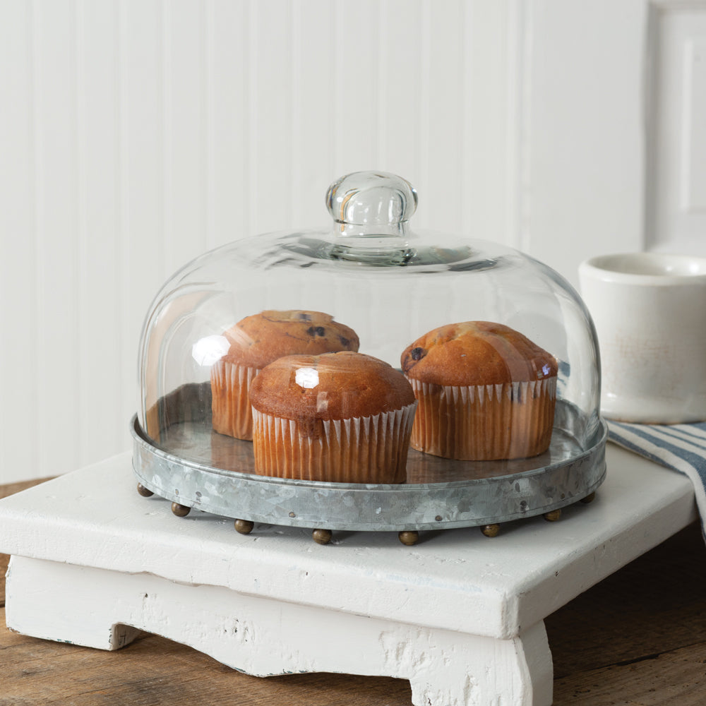 Glass cloche with brass-accented beaded base, showcasing elegant dessert display on a galvanized metal stand.