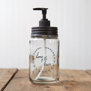 Clear glass soap dispenser with black pumper, featuring "Home of the Brave" and olive branches, perfect for patriotic decor.