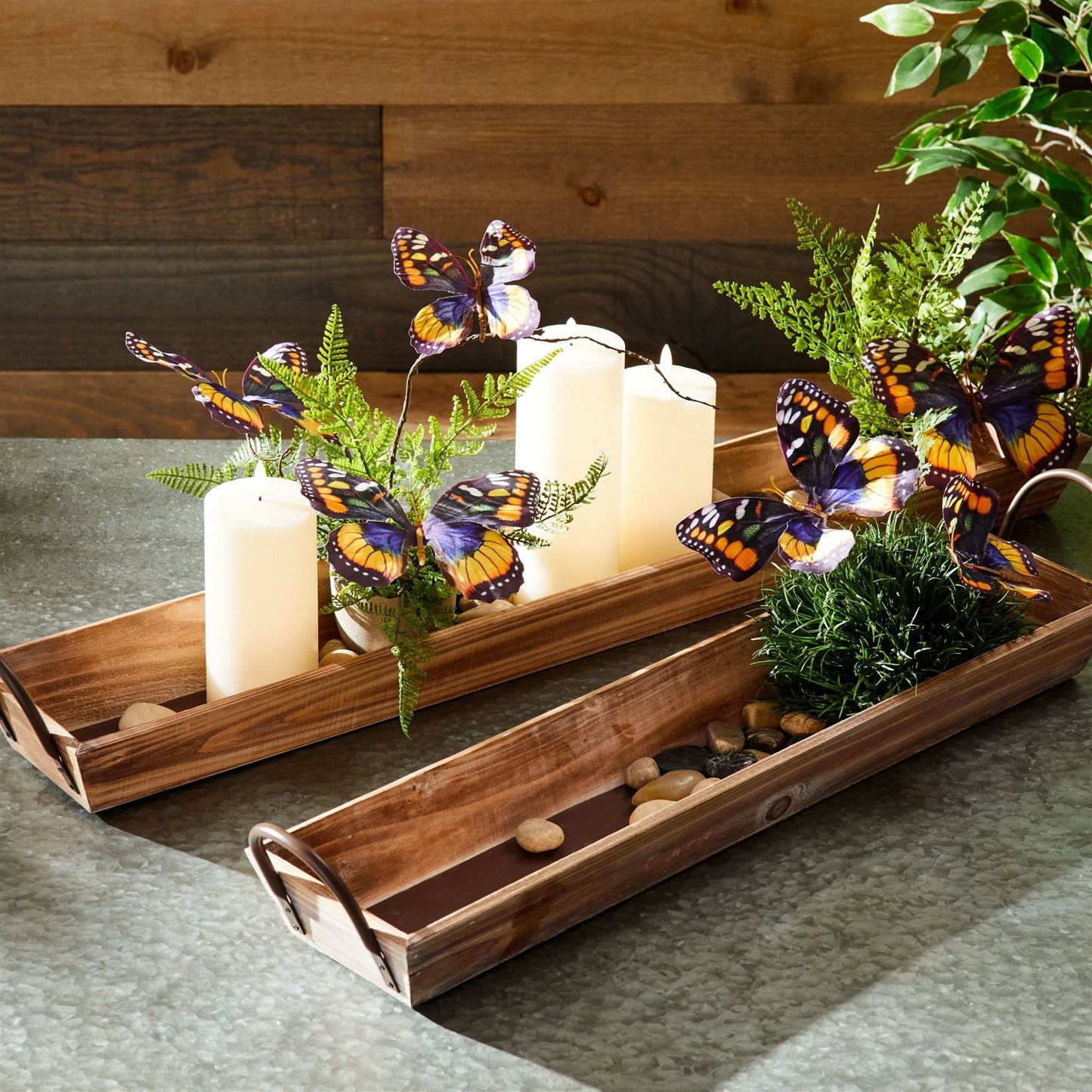 Decorative wooden trays with candles, butterflies, and plants on a wooden surface.