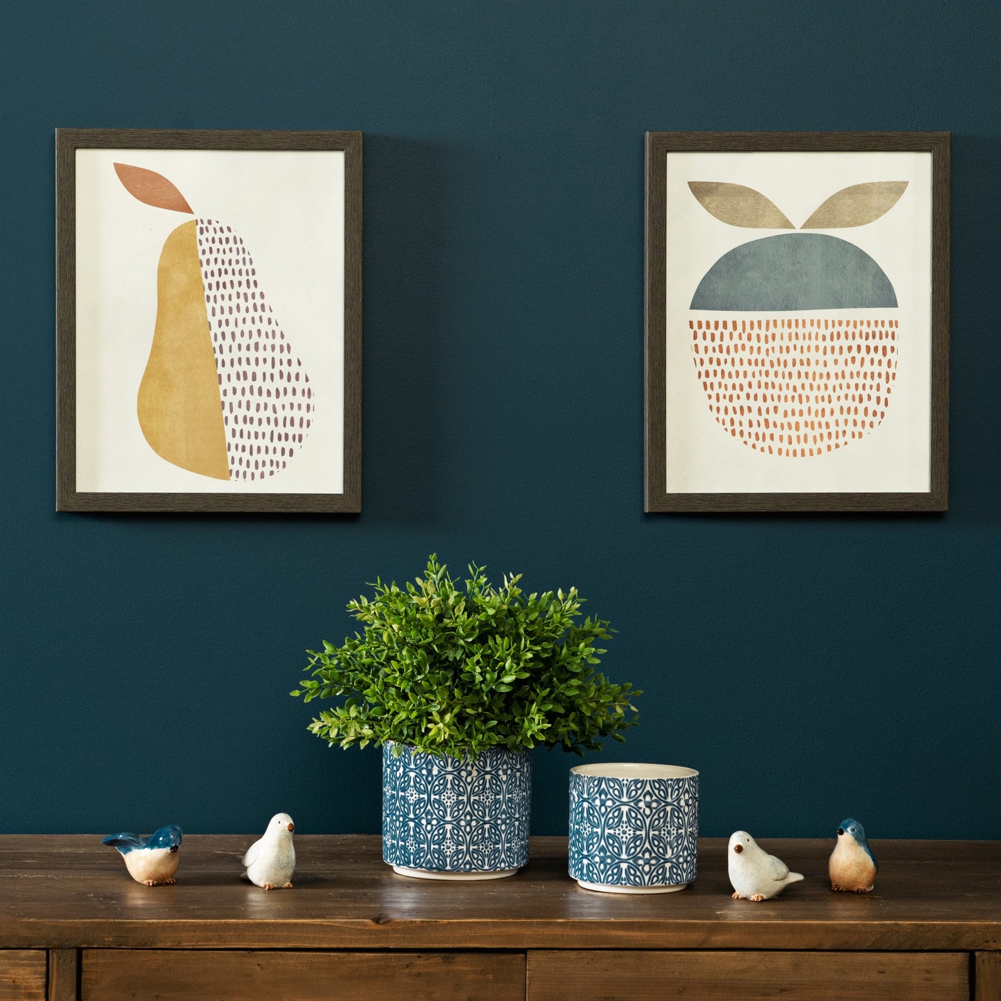 Two framed abstract artworks on a dark blue wall with a wooden shelf featuring decorative items.