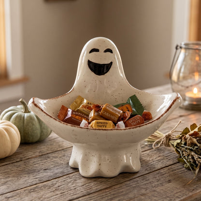 A ceramic ghost-shaped candy bowl filled with assorted Halloween candies, styled on a rustic wooden table with small pumpkins, dried grass, and a lit glass jar candle in the background.
