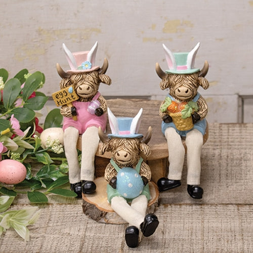 Highland cow figurines in pastel overalls and hats, with dangling fabric legs, holding Easter-themed items like eggs and carrots.