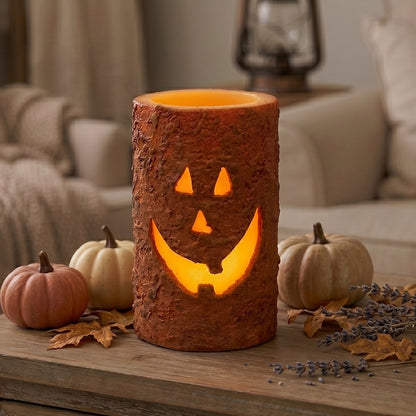 A textured faux log candle holder with a carved jack-o'-lantern face, glowing with warm orange light, displayed on a wooden table in a cozy living room setting with small pumpkins and fall leaves.