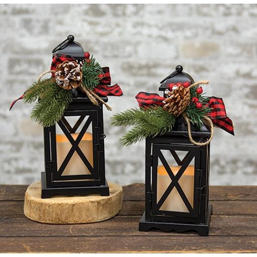 Two black metal lanterns with criss-cross panels, festive pine accents, and white LED candles casting a warm glow.