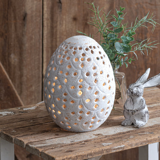 Rustic white perforated metal egg on a tabletop, perfect for decor. Elegant and versatile, ideal for adding charm to any space.